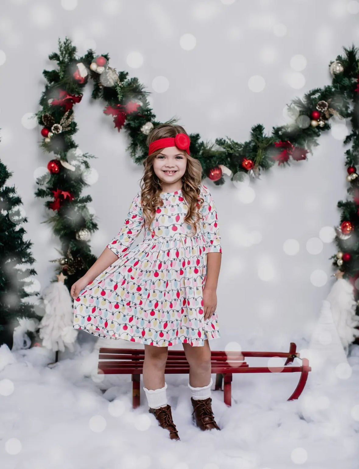 Girl wearing a Merry and Bright Pocket Twirl Dress, smiling in festive decor with a sleigh and holiday garland.